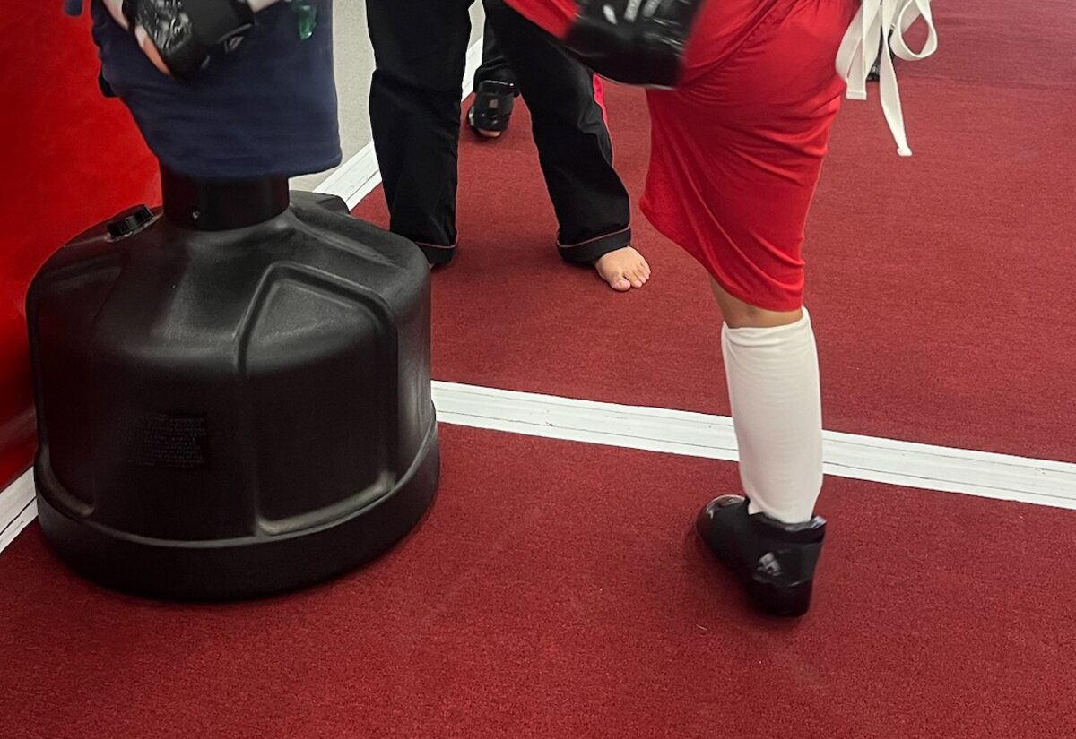 A student in shin guards kicks a freestanding bag as an instructor watches.