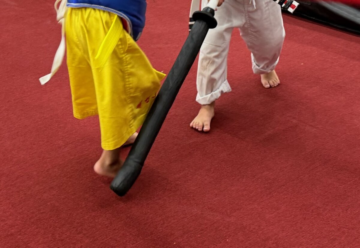 Two young students in uniforms practice with a foam sword on the red mat.