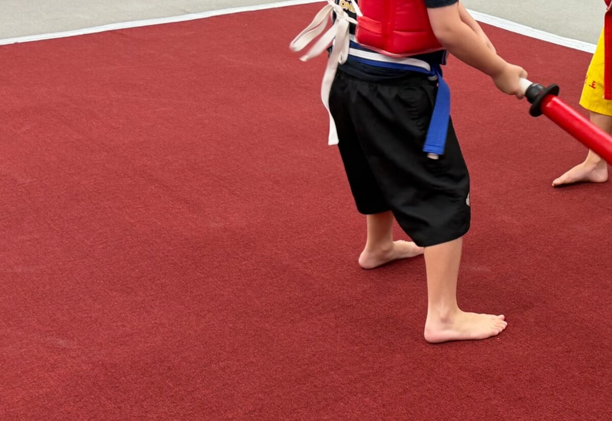 A student in a blue belt and chest protector holds a foam practice sword on the mat.
