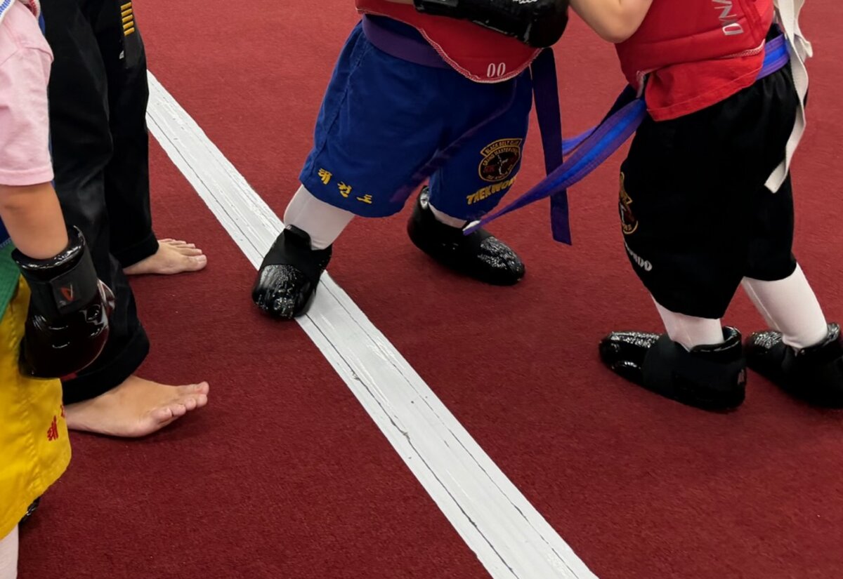 Students in chest protectors and foot pads wait at the white line beside barefoot classmates.