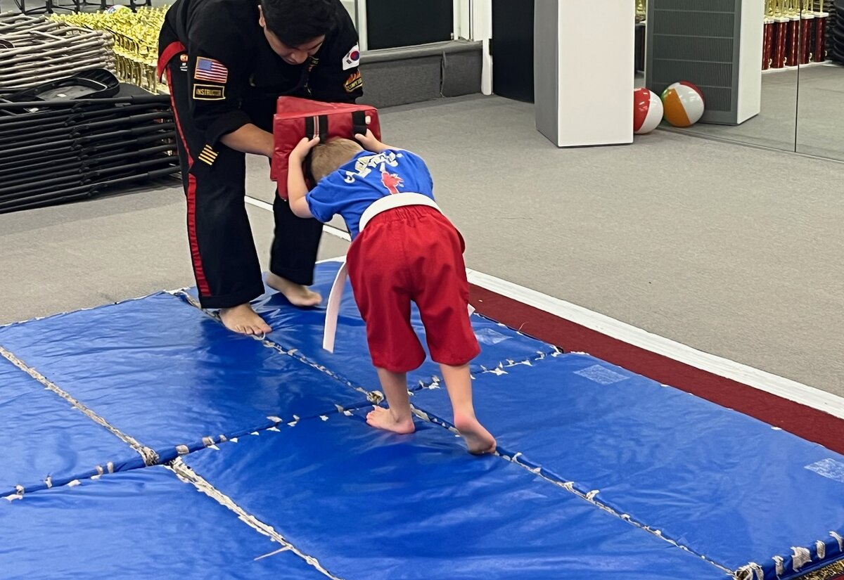 A young student in a white belt leans into a red pad that an instructor holds steady on the blue mats.