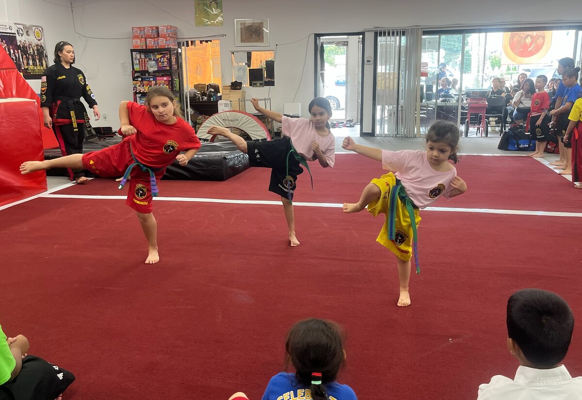 Three girls practice side kicks on the red mat while classmates sit at the edge and families watch from the lounge behind the glass.