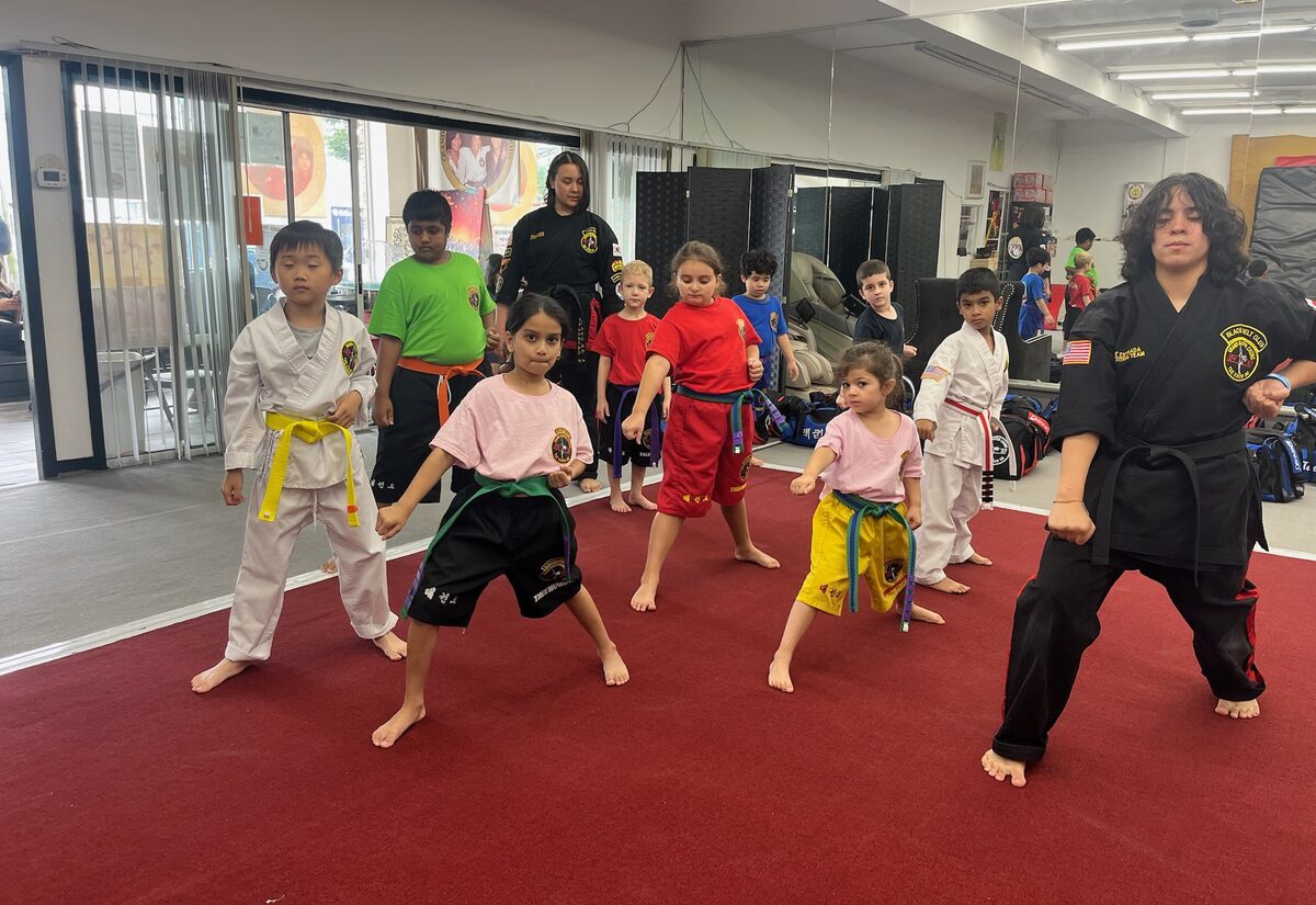Children hold a wide, low stance on the red mat, copying the black-belt instructor beside them.