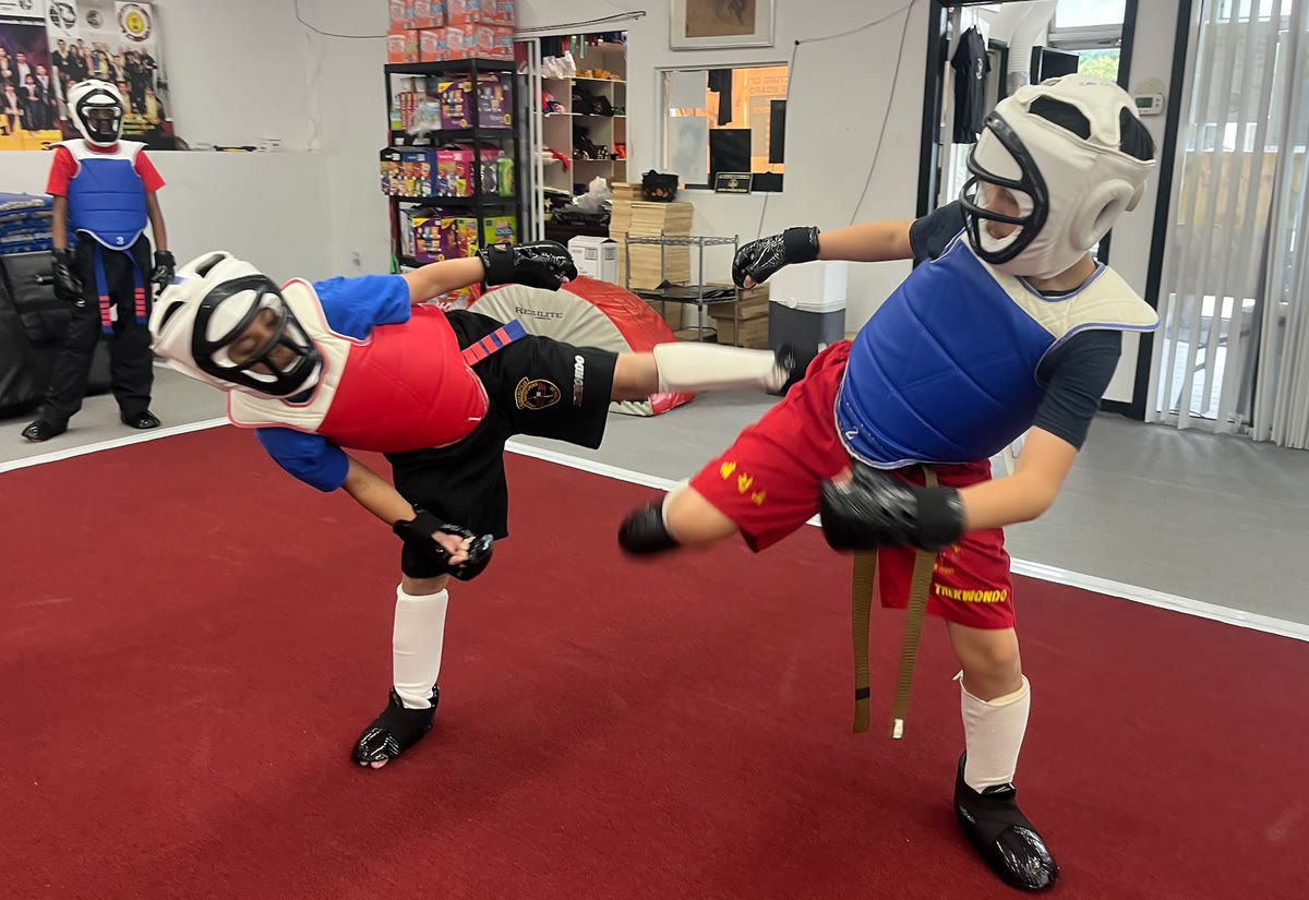 Two students in helmets, chest protectors and gloves spar on the red mat as another waits in gear behind them.