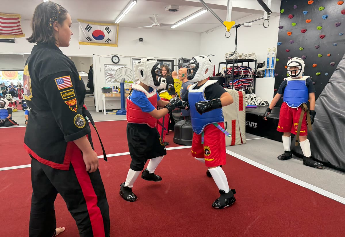 Two students in full sparring gear face off as a referee in a black uniform watches.