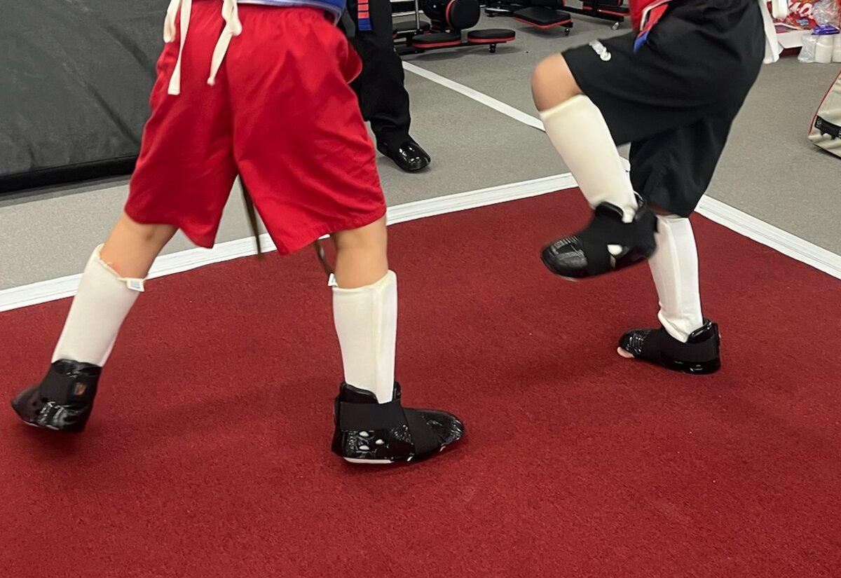 Two students in shin guards and foot pads move through a sparring drill on the red mat.