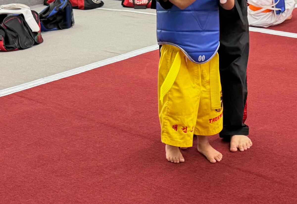 A young student in a yellow belt and blue chest protector stands on the red mat with an instructor close behind.